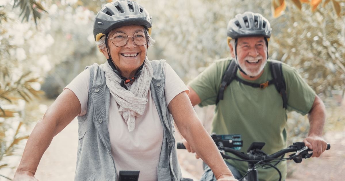 Smiling elderly couple riding bicycles outdoors wearing helmets, promoting wellness and health in MA.