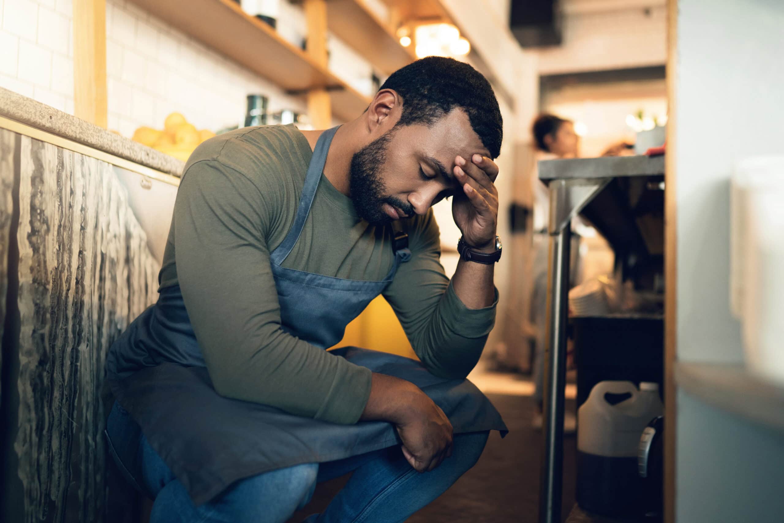 Exhausted man holding his head in a kitchen, feeling stressed or overwhelmed.