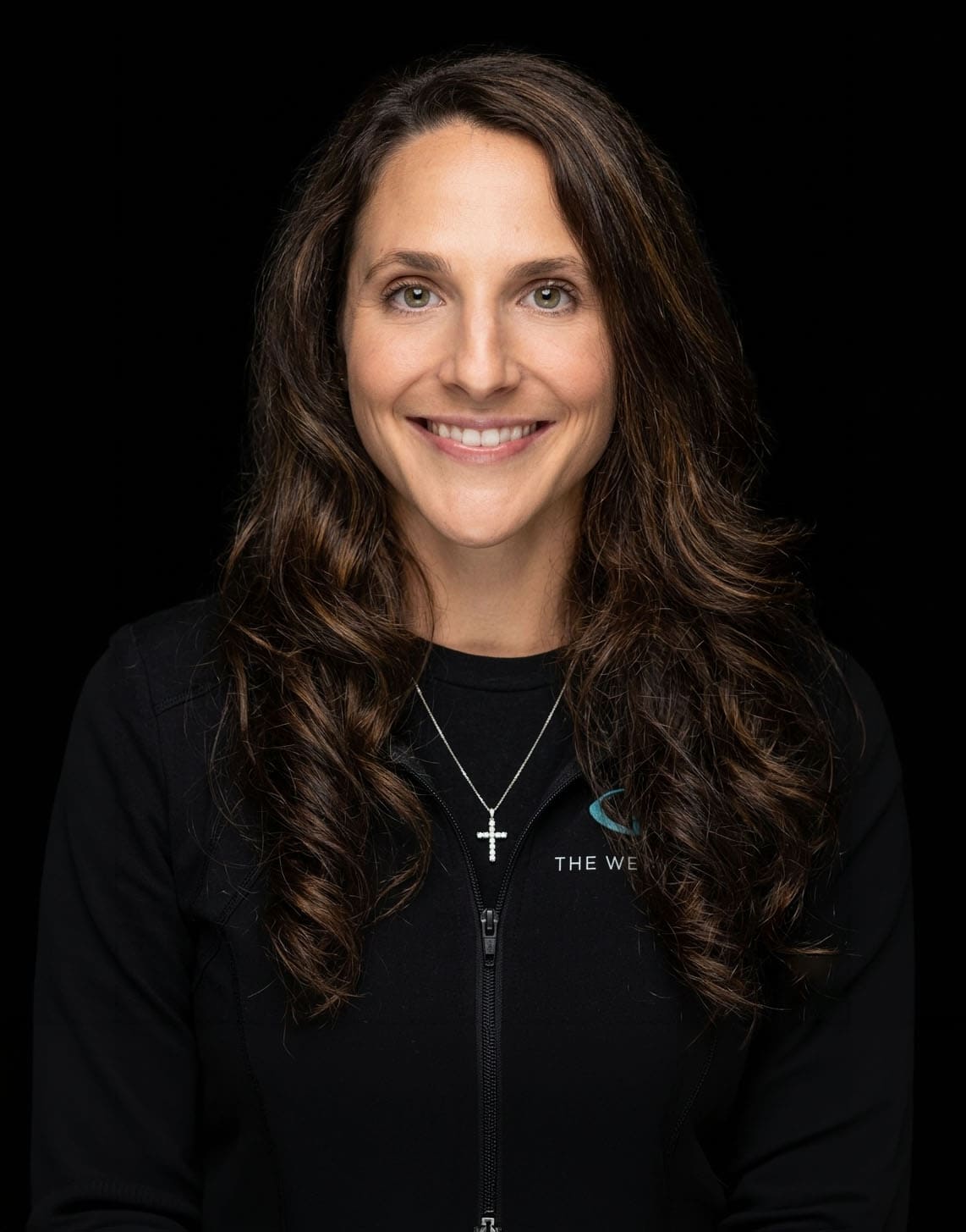 A friendly woman with long, wavy brown hair smiling, wearing a black jacket with "The Wellness Drip" logo, representing professional staff at Wellness Drip Holyoke.