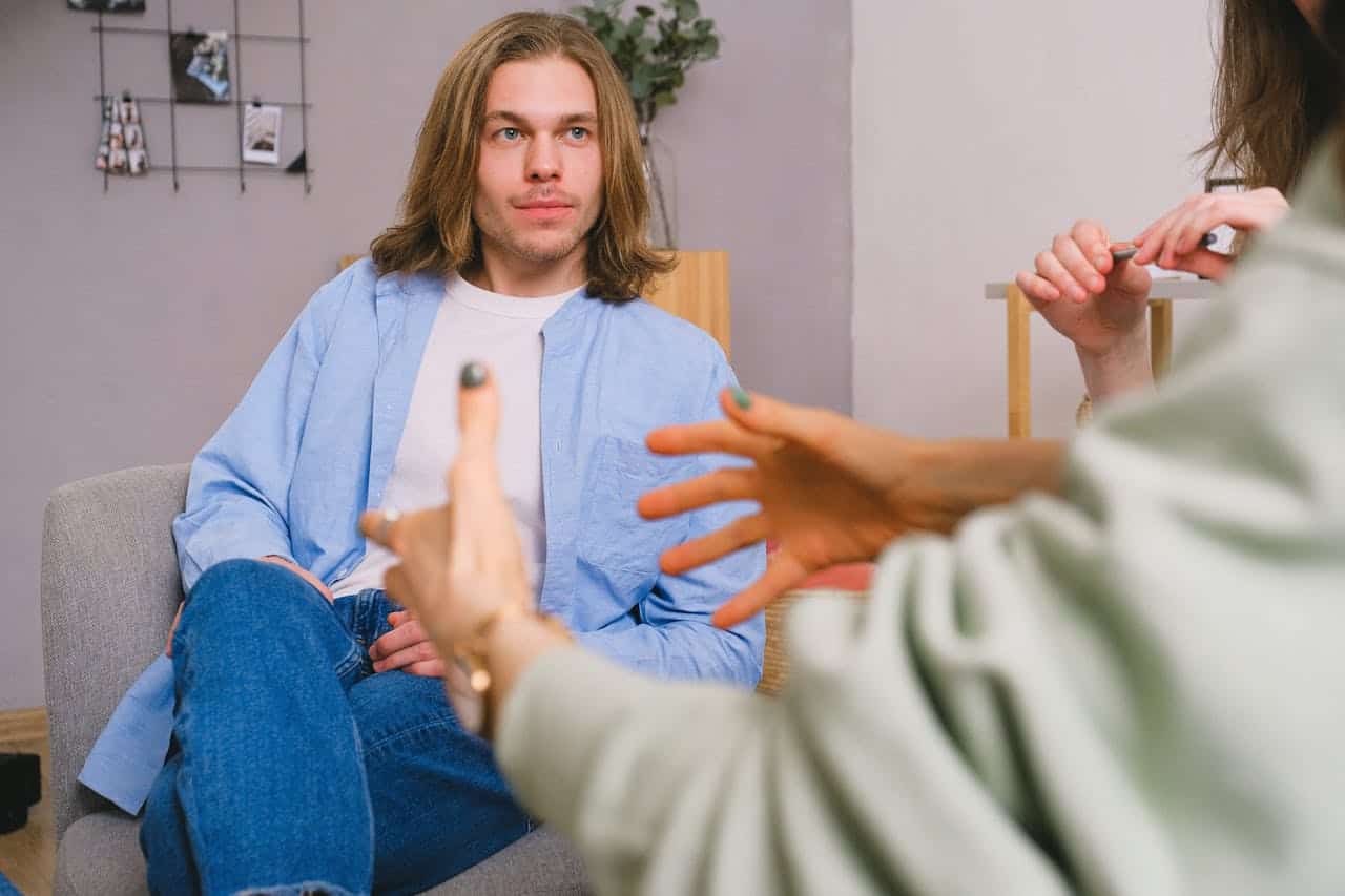 A young man with shoulder-length hair sitting on a sofa during an IV therapy consultation at Wellness Drip Holyoke, discussing health and wellness options with a healthcare professional.