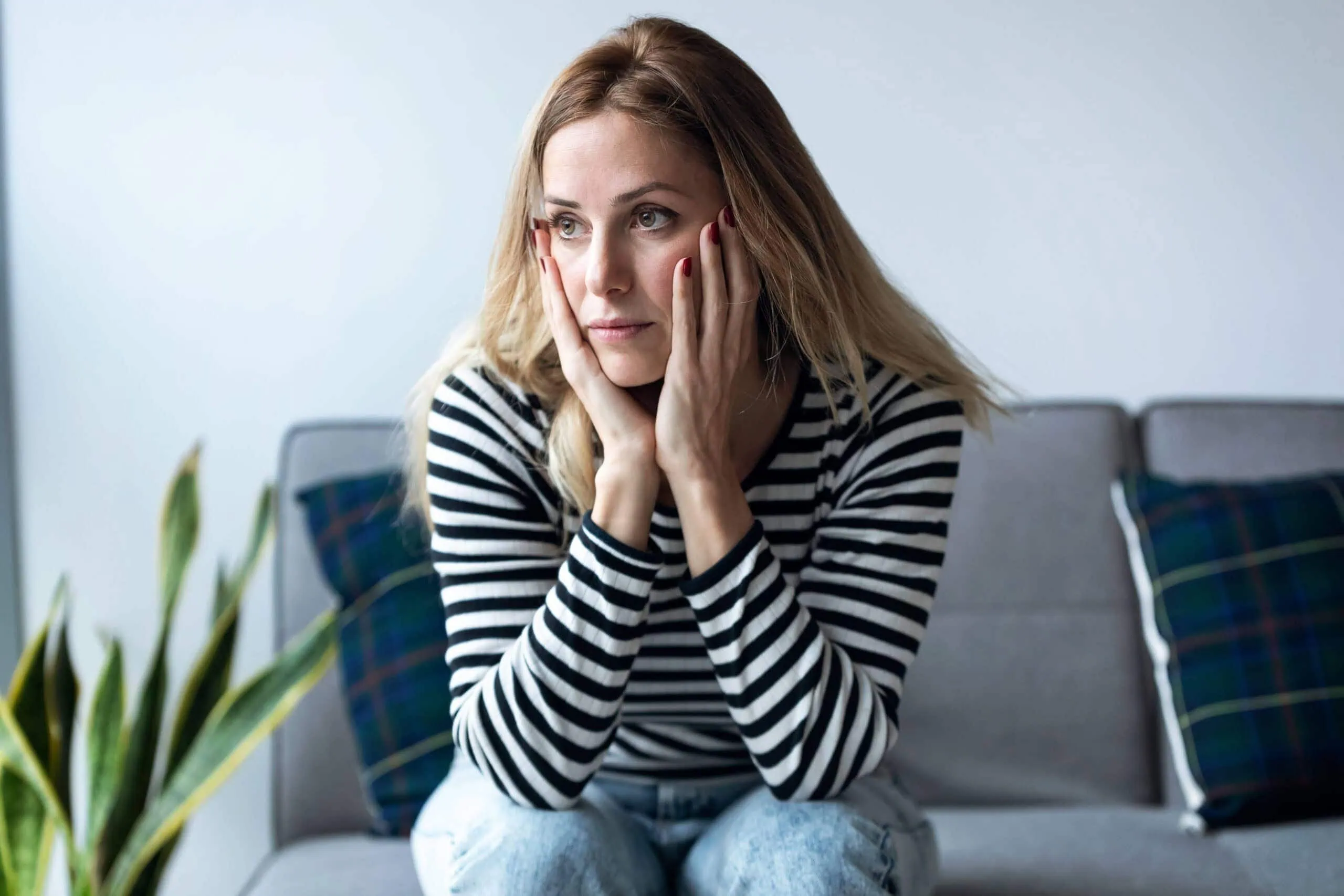 Anxious woman holding her face with hands, sitting on a gray sofa in a wellness clinic, experiencing stress or depression, mental health support, mood therapy, wellness services in Massachusetts.