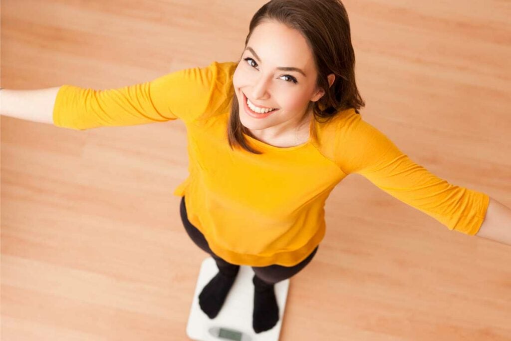 Bright young woman smiling on a scale, measuring weight and health progress at Wellness Drip INC, MA wellness clinic offering IV therapy, weight loss, and ketamine therapy services.