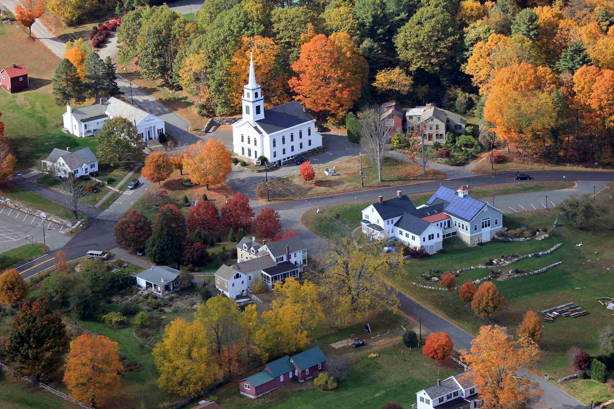 A picturesque aerial view of a small New England town during autumn, showcasing colorful fall foliage, charming houses, and a white church in a tranquil rural setting.
