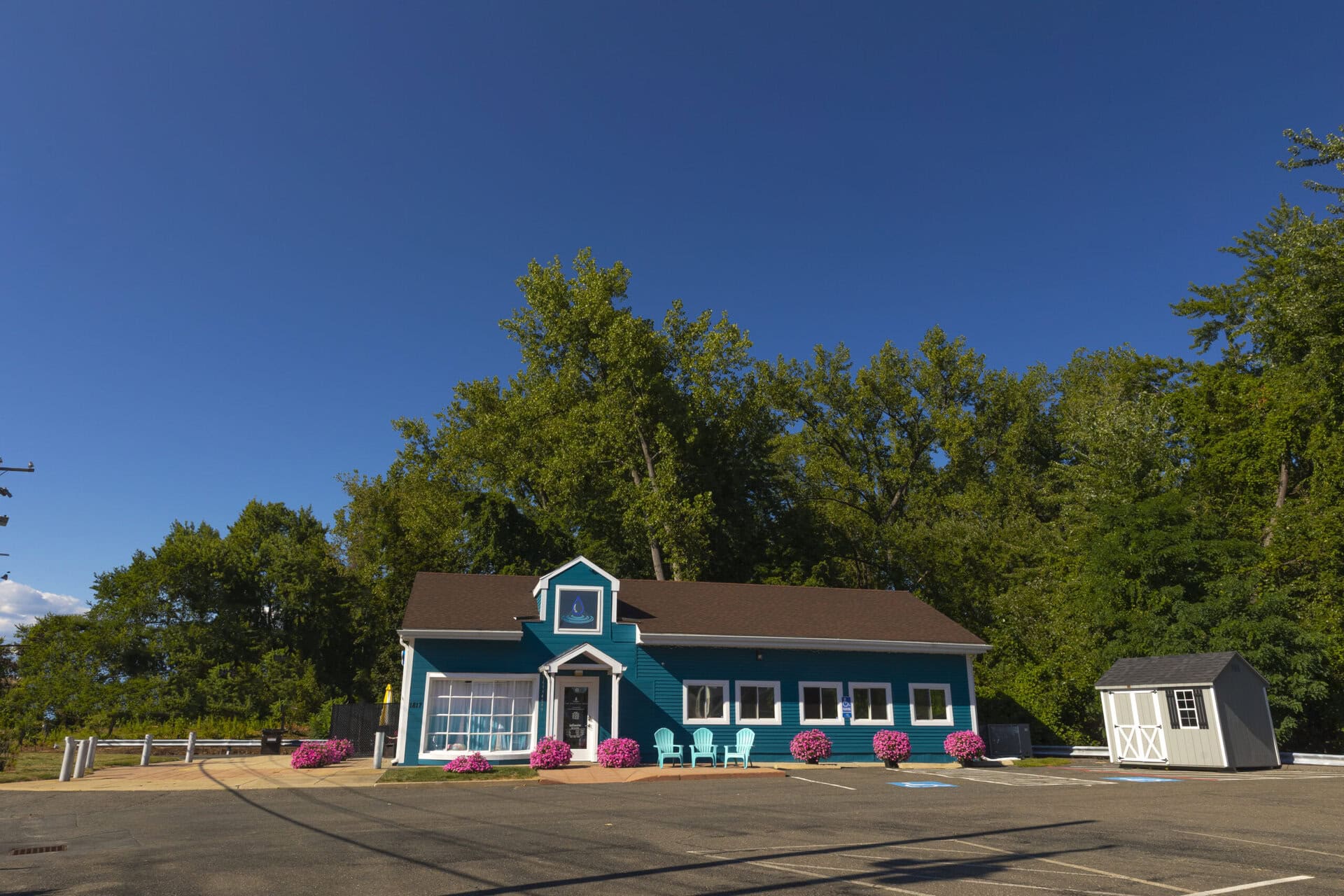 A charming blue wellness clinic with outdoor seating, blooming pink flowers, and a clear blue sky, specializing in health and wellness treatments.
