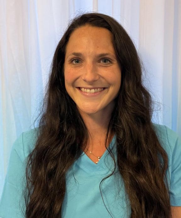 A smiling woman with long dark hair wearing a light blue shirt, standing in front of a white curtain, representing the friendly and professional staff at Wellness Drip INC.