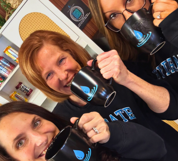 Group of women at Wellness Drip INC smiling and holding branded mugs during hydration therapy session at the wellness clinic.
